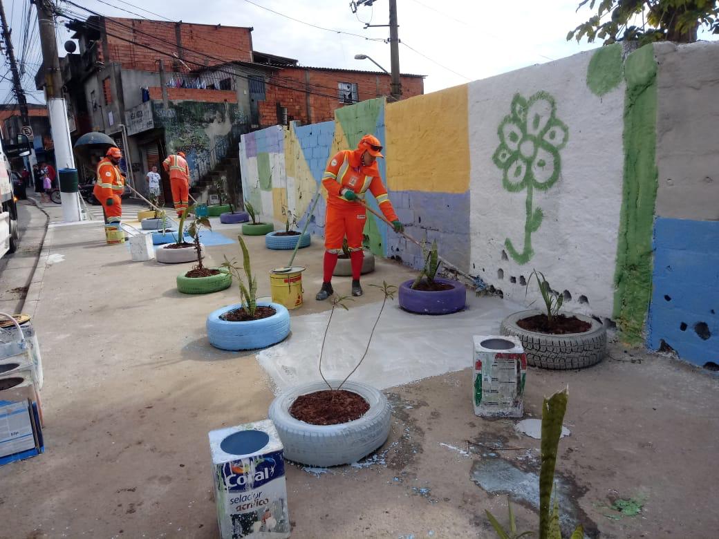 Quatro homens com uniforme laranja pintam calçada e pneus que serão usados como floreiras